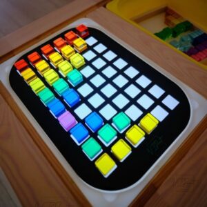 A brightly colored grid featuring various transparent and colored blocks arranged in a rainbow pattern on a black background, illuminated from below. The blocks are arranged in rows and columns with some empty spaces, along with additional multicolored blocks visible in a compartment nearby.