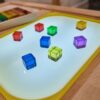 A light table made from IKEA Flisat sensory table and VIPrint studio light table insert, with colorful transparent acrylic cubes in various shapes and sizes arranged on it, set against a smooth yellow border, which is feature of IKEA trofast bin, providing a sensory play experience ideal for toddlers.
