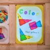 A light table made from IKEA Flisat sensory table and VIPrint Studio light table insert, showing colorful translucent triangles in various colors, with the word 'Color' handwritten above and a question 'blue + yellow = ?' below, surrounded by bins of similar shapes, which creates a sensory play idea for toddlers and early introduction of mixing the light colors.