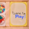 An overhead view of an Ikea Flisat sensory table featuring a VIPrint Studio light table insert with the text 'time to Play!' displayed in colorful letters, alongside two IKEA trofast bins containing transparent and colorful geometric shapes of magnetic tiles.