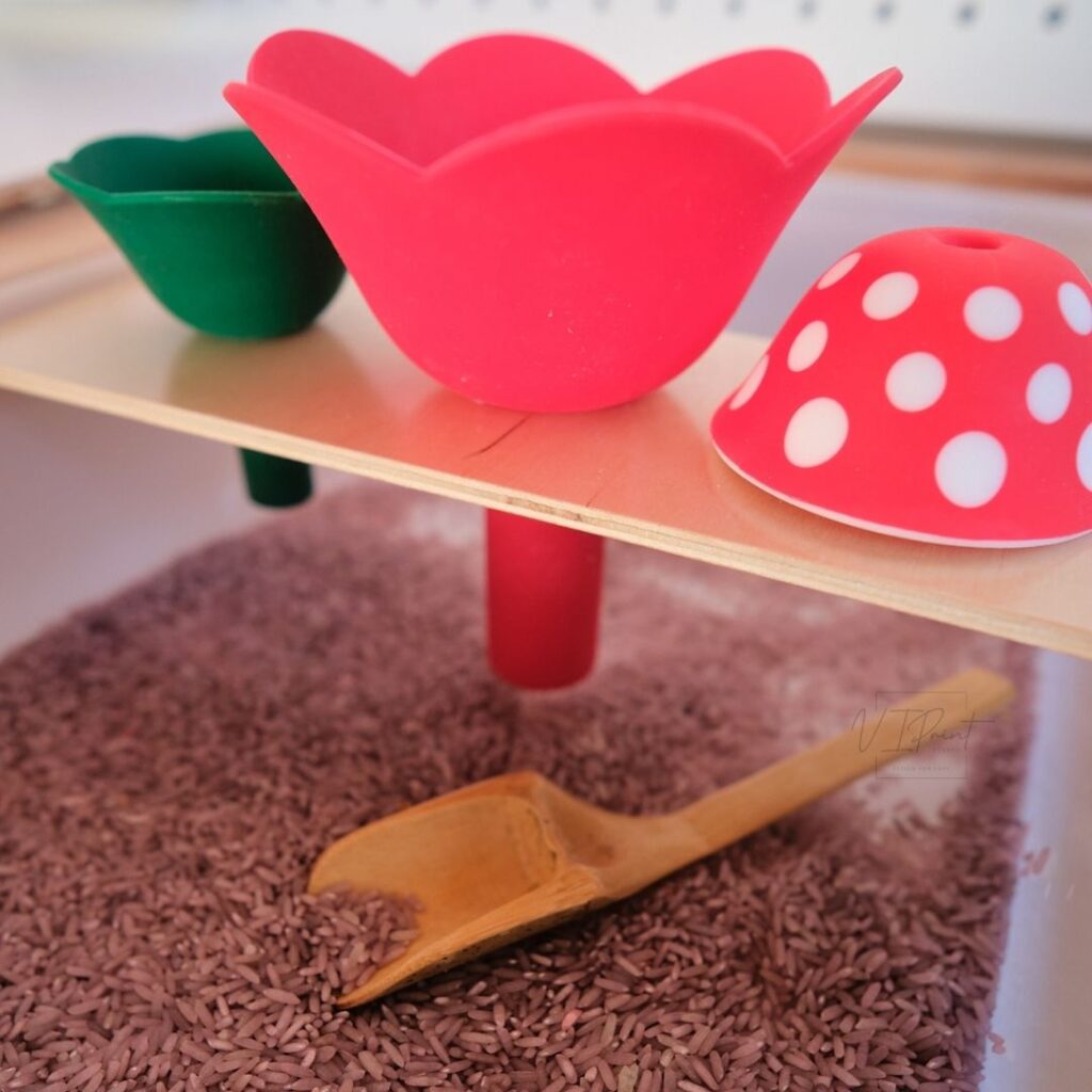 A set up of toddler activity of colorful ikea flisat sensory table featuring a variety of silicone funnels in red and green on a wooden board above an ikea trofast bin filled with purple-colored rice, with a wooden scoop placed nearby.